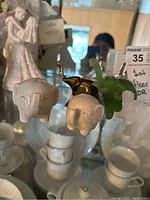 Four small decorative elephant figurines: two white ceramic with pink details, one brass, and one green glass. Shown on a reflective surface with some background objects.