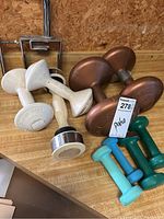Various free weights arranged on a wooden surface including white cast iron dumbbells, brown round dumbbells, and smaller colored dumbbells.