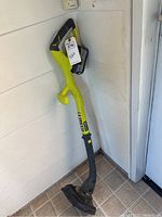 Single image showing a green and black Ryobi cordless weedwacker trimmer with no battery or charger, standing against a white wall and tiled floor.