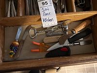 Photo of a drawer compartment containing various cutting tools and kitchen utensils including a corkscrew, paring knives, vegetable peeler, and hand-held cheese slicer.