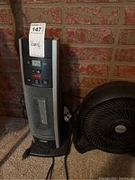 Vertical space heater next to a small black fan against a brick wall.