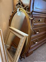 Two wall mirrors leaning against a dresser, larger ornate oval gold-framed mirror in back, smaller rectangular cream-framed mirror in front
