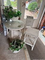 Full view of the wicker furniture set placed inside near glass doors, showing the round glass top side table, square side table, tall plant stand, and two flower pots with greenery.