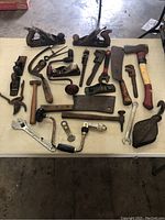 Full view of all vintage hand tools arranged on a table including wood planes, hammers, wrenches, hand drills, axes, cleaver, and pulley.
