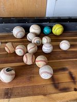 Overview of 14 balls spread on wooden floor: mostly baseballs, one yellow softball, one turquoise ball.