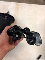 Close-up of Tasco binoculars showing model 7x50 and serial number, held in hand, showing front lenses and focus wheel.