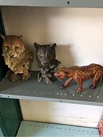 Three tiger figures displayed on a metal shelf: large tiger bust, black tiger bust, and walking cheetah.