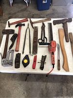 Overview of assorted used hand and power tools arranged on white table surface, including hammers, pliers, scissors, tape measure, and electric hedge trimmer.