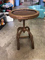 Full view of the vintage adjustable stool showing the wooden seat and iron legs on a concrete floor in a garage or storage area.