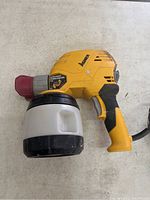 Side view of yellow Wagner Power Painter Plus 6.6 paint sprayer with attached paint container on beige surface.