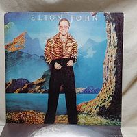 Front cover of Elton John's 'Caribou' album showing him in a tiger-striped jacket against a painted mountain backdrop.