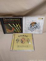 Three music CDs placed on a surface: Lovin' Spoonful Greatest Hits, Red Mann's Earth Band album, and Tonic Lemon Parade album.