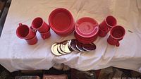 Eight red ceramic Fiesta coffee mugs arranged in pairs, eight red salad plates stacked, six red bowls stacked, and seven shiny chrome condiment trays laid out on a white cloth background.
