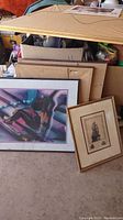 Photo showing two framed art pieces: a colorful reclining figure artwork and a vintage ship print leaning in front of several other framed artworks and boxes.