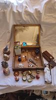 Wooden box with attached mirror on lid holding various small perfume bottles and trinket boxes around it, plus a porcelain doll wrapped in cloth.