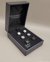 Open black DiamonLuxe box showing three pairs of sterling silver earrings: pearl studs, clear square simulated zirconia studs, and black square simulated zirconia studs.