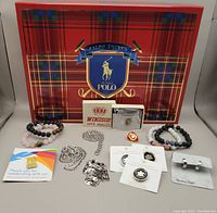 Full lot arranged on Ralph Lauren Polo box showing various jewellery items including stone stretch bracelets, stainless steel chains, pins and new lighter in box.