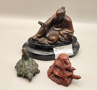 Three Asian figurines including metal scholar figurine on wooden base, bronze man riding turtle figurine, and wooden stacked turtles figurine photographed together on a flat surface.