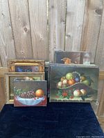 Ten signed canvas paintings stacked and leaning on fence, including some gold-framed 8x10s, various fruit still life scenes and a dog painting visible.