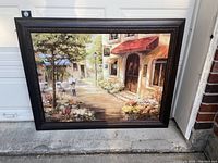 Full view of large framed canvas painting showing a charming street scene with flowers, trees, and a building with a red awning.