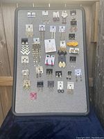 Photo shows a display board with 32 pairs of assorted dangle and drop style pierced costume earrings arranged in rows, showcasing varied shapes, sizes and colors.