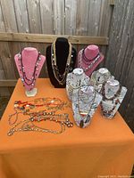 Full lot view of 26 costume necklaces displayed on various busts and orange table, showing assortment of gold and silver tone beads and rhinestones.