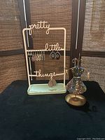 Metal jewelry stand with 'pretty little things' text holding assorted earrings and rhinestone dress jewelry holder on black cloth backdrop.