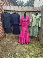 Four evening attire outfits on mannequins displayed outdoors against wooden fence including a bright fuchsia dress and jacket, navy-blue skirt set, and two other skirt sets.