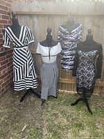 Four mannequins displaying the black and white dresses and outfit outdoors against a wooden fence, showcasing different patterns including stripes and chain print.