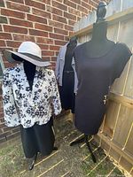 Shows the black and white floral printed blazer, black dress, striped jacket and black and white floppy hat on mannequins against a brick wall and wooden fence.