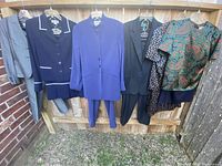 Full view of six hanging ladies clothing sets on hangers in outdoor setting, including mainly blue and gray dresses, pantsuits, and tops, showing variety of cuts and styles.