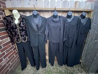 Full view of all pantsuits and tops displayed on mannequins hanging on a wooden rack outdoors against a brick wall and fence.