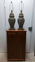 Closed wood cabinet with two metal lamps placed on top. Cabinet has double doors with knobs, lamps have ornate cut-out designs with harps and finials.