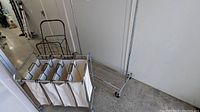 Photo showing collapsible garment rack with wheels, 4-section fabric hamper attached, and folding shopping cart nearby on concrete floor against white wall.