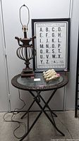 Full view of lamp, table with glass top and woven edge, phonetic alphabet art behind, and floral posts on table surface.