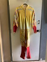 Full front view of yellow and red ski jumpsuit with wing and flame graphics hanging on a door.