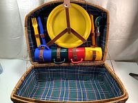 Open picnic basket showing 4 yellow plastic plates, fork, knife, spoon, and 4 multicolored cups secured with leather straps inside lid with plaid fabric lining.