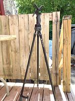 The full view of the Primos Trigger Stick GEN 3 Jim Shockey Edition tall tripod standing on a wooden deck, showing its three legs fully extended and V-shaped padded yoke on top.