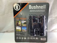 Front view of sealed Bushnell 10x42 binoculars in original packaging, showing product and usage icons.