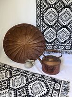 View of two black and white geometric patterned cotton floor mats, handmade pottery jug, vintage copper kettle, and large hand-carved wooden tray arranged together
