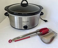 Crock-Pot slow cooker with stainless steel body, black ceramic insert, glass lid, red-handled tongs, and metal double spoon rest