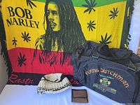Large colorful Bob Marley blanket displayed as backdrop with wool toque, wallet and black duffle bag in front