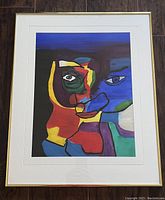 Photo of the original abstract painting showing the full artwork in a gold metal frame with white mat border. The painting illustrates a vividly colored abstract face.