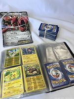 Photo showing a binder open with several Pokemon cards visible including Pikachu and Scizor EX, a stack of loose cards tied with a rubber band, and some cards displayed outside the binder.