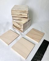 Photo showing two stacks of sealed and shrink-wrapped sets of 24 unfinished pine square coasters with three individual coasters and a strip of round black foam feet pads laid in front.