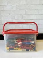 Front and side view of transparent plastic bin filled with assorted vintage Lego bricks, with red handle and lid.