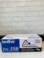 Front view of sealed Brother TN-350 toner cartridge box against a white brick wall background.