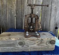 Front view of a vintage metal pipe clamp mounted on a thick weathered wooden block, showing the turning handle, lever, and bolts attaching clamp to wood.