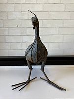 Front view of the bird sculpture showing its rounded riveted body and long, slender legs with splayed toes.