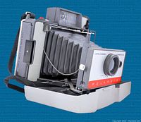 Front and right side view of vintage Polaroid Land Camera 104 showing bellows, lens, Polaroid branding and attached strap with protective plastic base case.